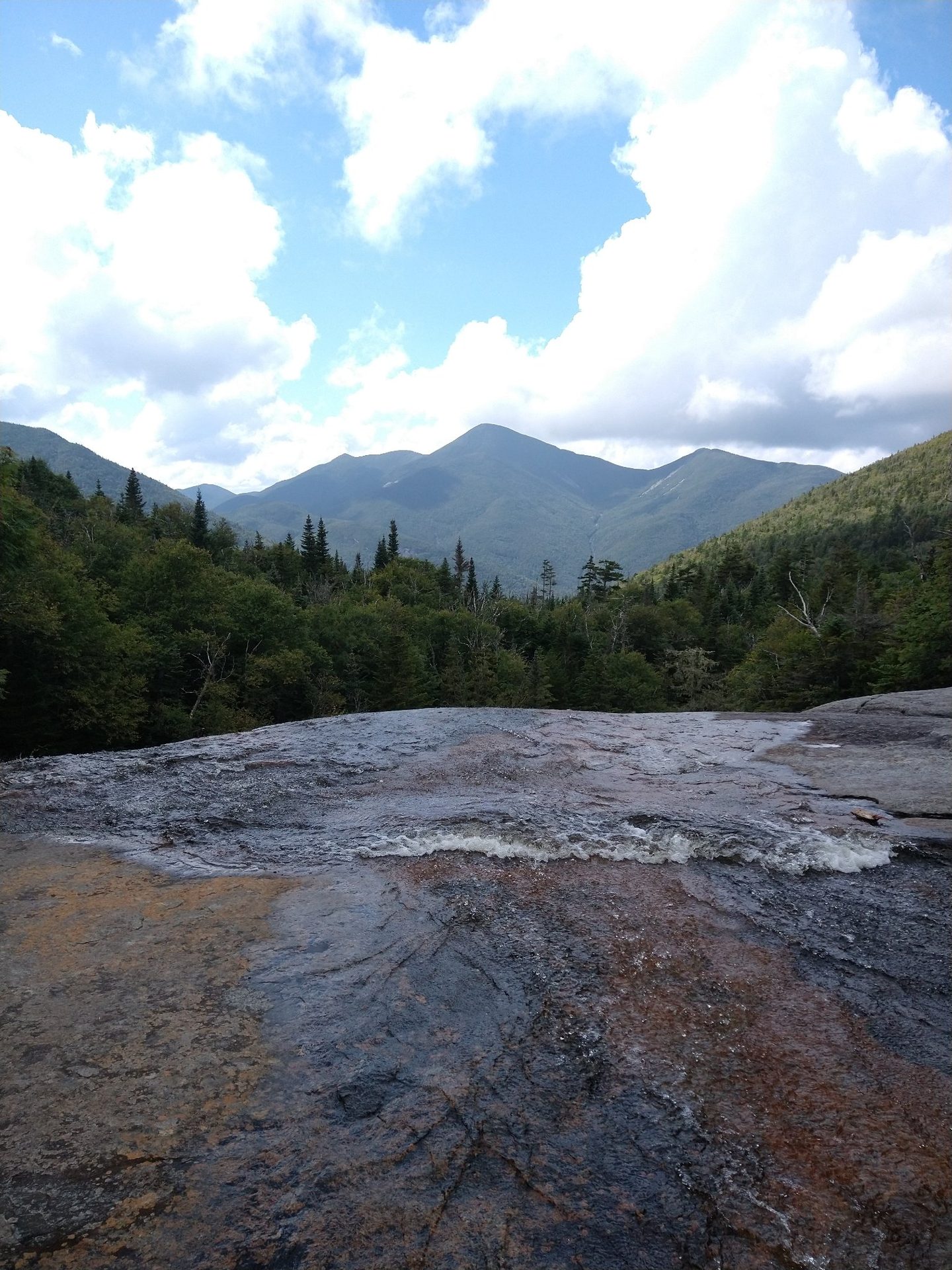 A high mountain peak seen across a wet rocky slab and forest