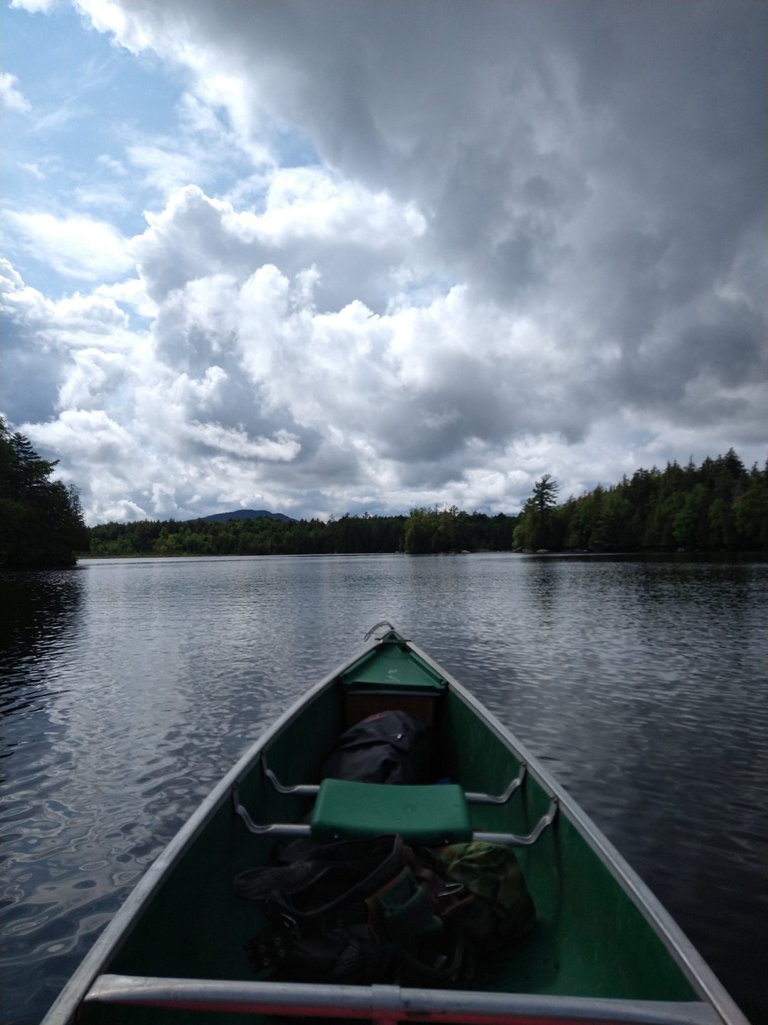 View from the bow of a green canoe crossing a calm Adirondack lake under dramatic clouds